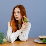 Caucasian woman sitting at table holding lime with broccoli and melon.